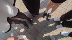 A group of people stands in a circle on a sunlit paved surface. Only their feet and legs are visible, showcasing a variety of stylish footwear, including black heels, loafers, and white sneakers. The ground features a patterned layout of gray and brown bricks.