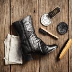 A black leather boot sits on a wooden surface, accompanied by an array of shoe polishing tools. Among them are two types of shoe brushes essential for maintaining shine, alongside a soft cloth and tins of white and black polish.